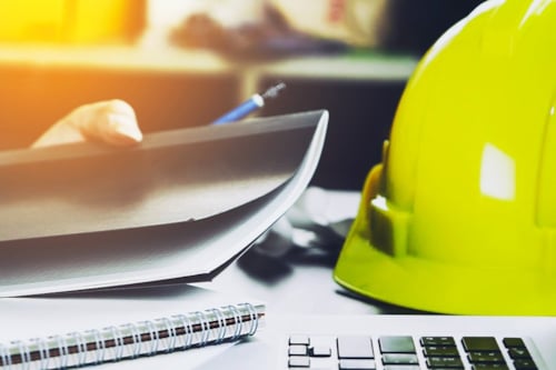 En gul bygghjälm ligger på ett skrivbord bredvid en dator och pappersdokument.