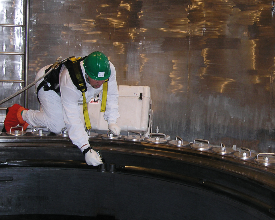 Person i skyddsdräkt och hjälm inspekterar industriell utrustning i en metallisk miljö.