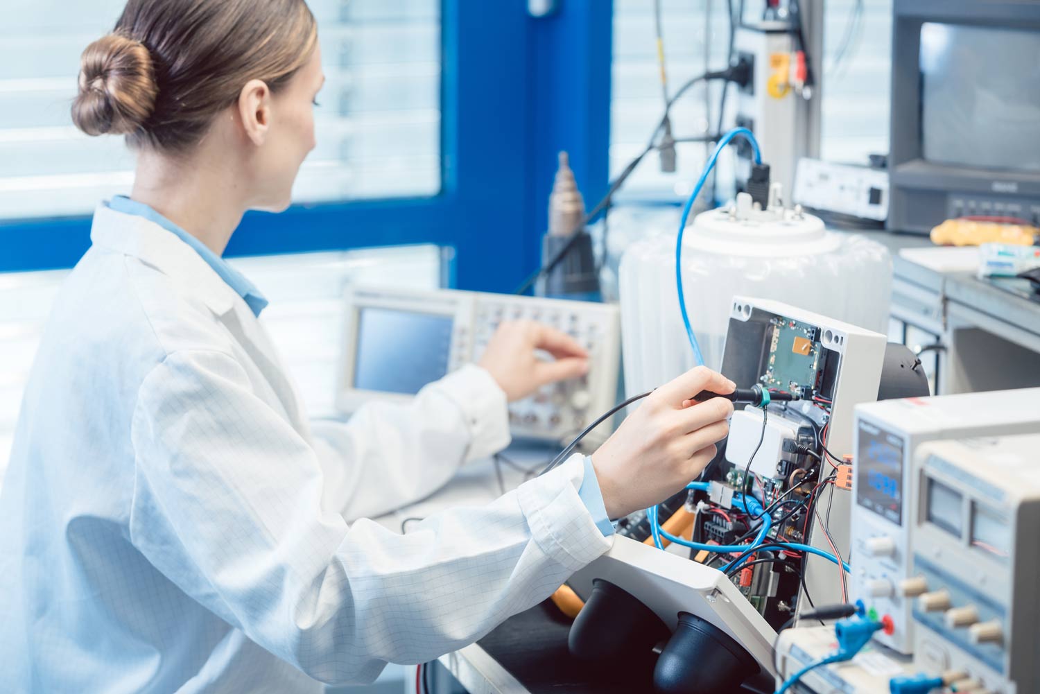 A female Kiwa lab employee in a white lab coat works on electronic equipment in a laboratory, using measurement tools and oscilloscopes for diagnostics and testing.