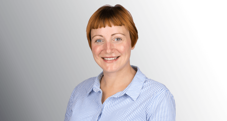 A smiling Gemma Ufton with short red hair wearing a blue and white striped shirt, set against a light grey background