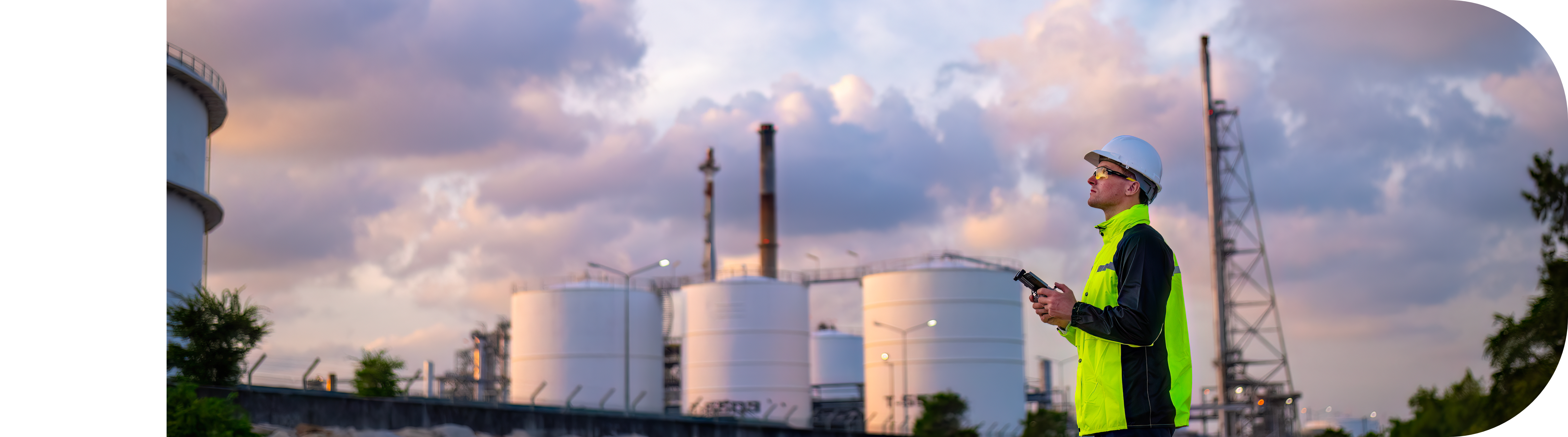 Industrial engineer in high-visibility clothing and hard hat operates equipment at an oil refinery with storage tanks and smokestacks in the background during a cloudy sunset