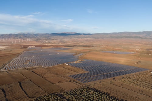 Vista aérea de un extenso parque solar en un paisaje rural de España, con montañas al fondo y un campo de aerogeneradores a la izquierda.
