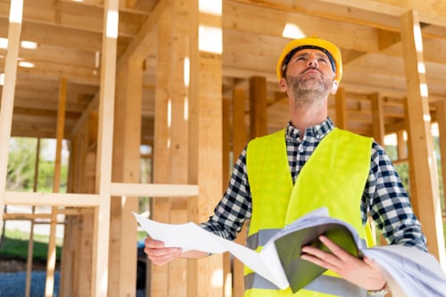 Manlig byggprojektledare i skyddshjälm och varselväst står med dokument i händerna framför en byggkonstruktion i trä.