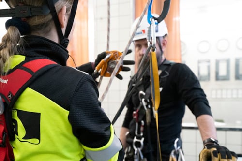 Två personer i skyddsutrustning tränar på arbetssäkerhet med rep och selar inomhus.